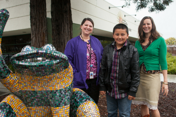 two professionals and a boy stand next to frog scupture outside dental campus